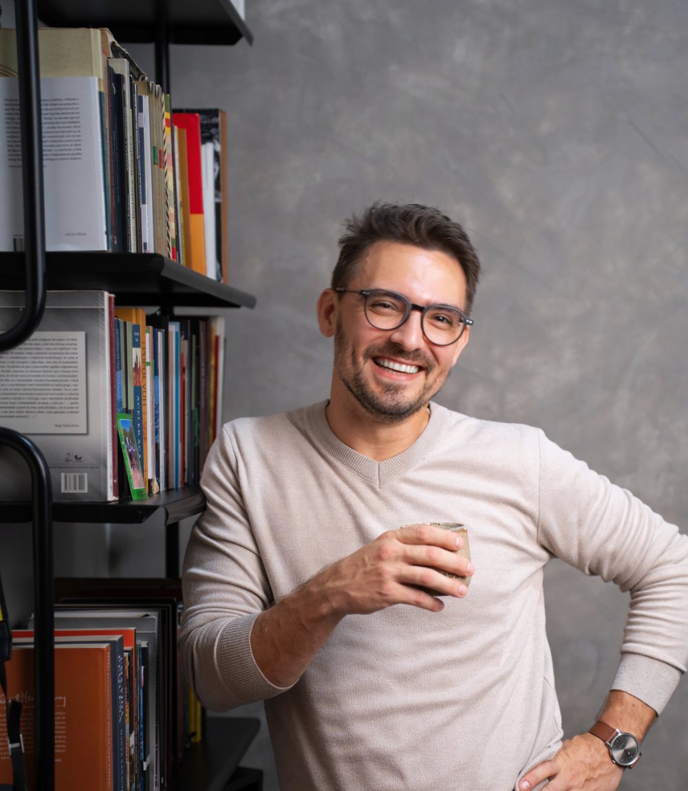 Rodrigo A. B. Pereira, psicólogo, de pé ao lado da estante de livros do consultório, com uma xícara de café na mão
